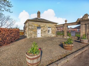 A stone building with a white door, a gravel driveway, and two wooden barrels with plants. - South Lodge - UK48318 (Berwick-Upon-Tweed)