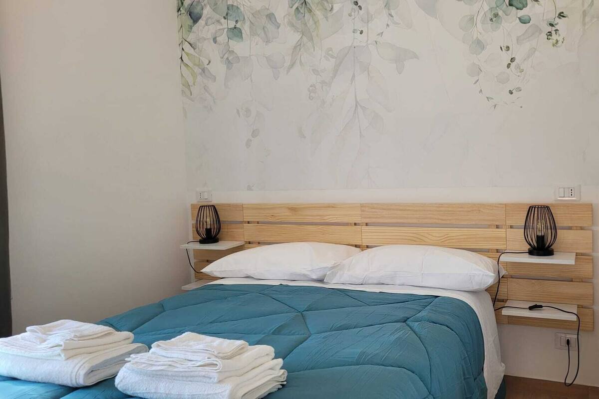 Un dormitorio con cabecera de madera, almohadas blancas, colcha azul turquesa y un mural en la pared.