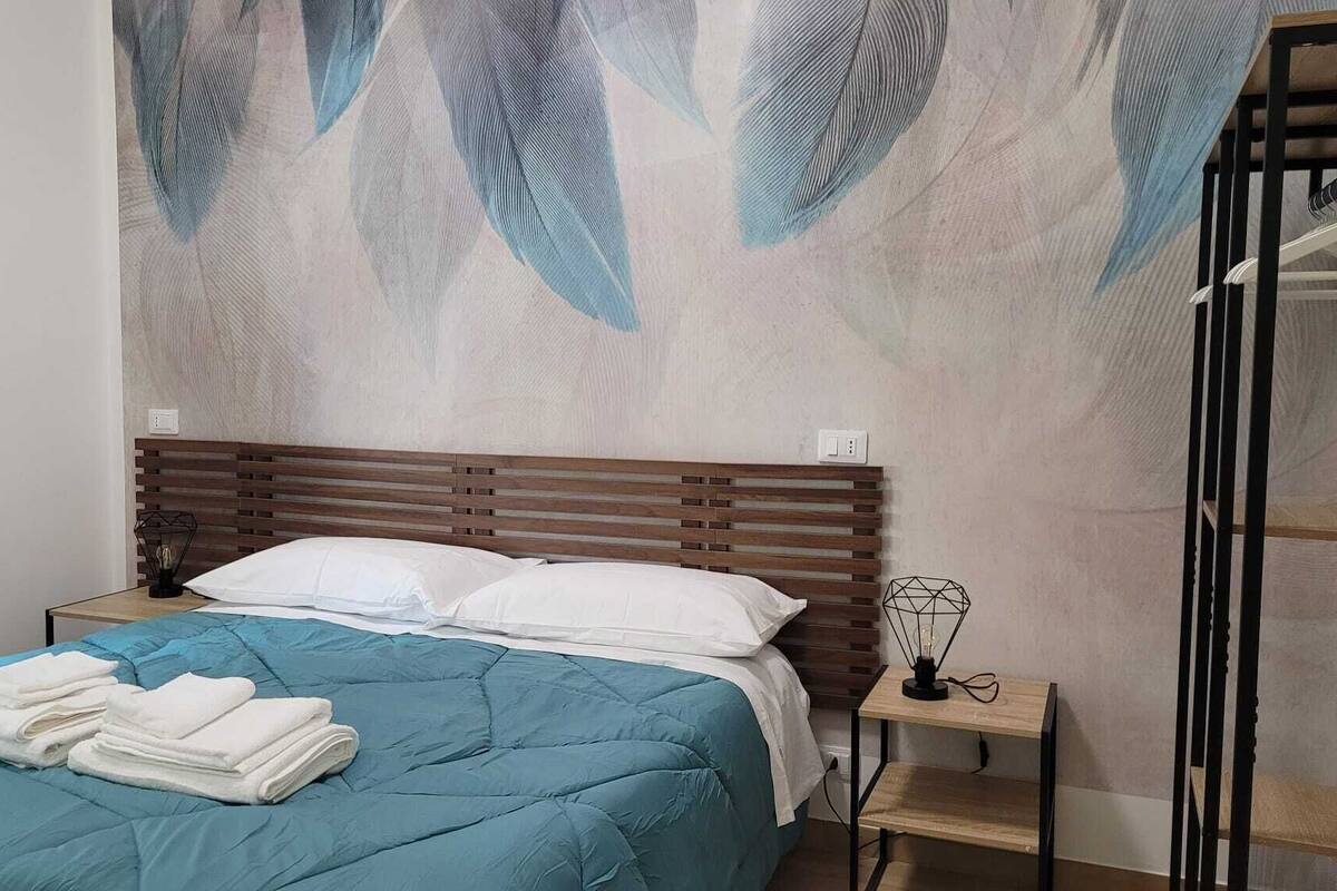 Un dormitorio con cama, mesita de noche, pared con diseño de plumas y estante con lámpara.