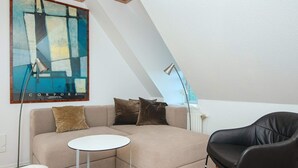 Um quarto de loft moderno com sofá seccional, uma mesa redonda pequena e uma poltrona de couro preta. Uma grande pintura abstrata decora a parede.