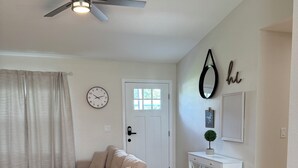 Una sala de estar con un sofá seccional color beige, un ventilador de techo, una puerta blanca, un reloj de pared y una pequeña consola blanca con una planta.