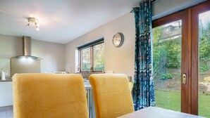 A dining area with a wooden table and yellow chairs, a kitchen in the background, a clock on the wall, and a view of a garden through a glass door. - Beautiful 3-Bed Riverside Townhouse with Parking - Pass the Keys (Belfast)