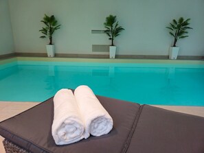 A poolside area with a lounge chair and two rolled towels. There are three potted plants on the wall.