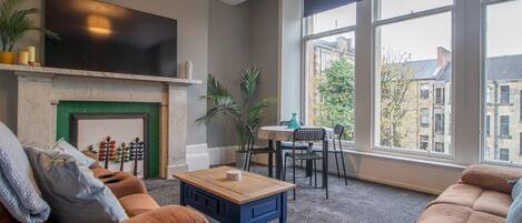 A living room with a fireplace, a television mounted above it, a dining area with a table and chairs, and large windows offering a view of buildings outside.