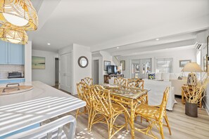 A modern kitchen with white cabinets, a built-in oven, and a dining area with wicker chairs and a glass table. - Radiant Goose Rocks Beach Getaway (Kennebunkport)