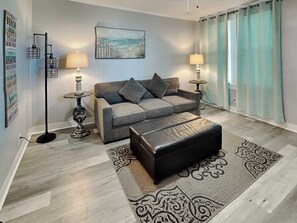 Una sala de estar moderna con un sofá gris, dos lámparas de pie, un tapete estampado y un cuadro con una playa.