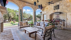 A stone-walled patio with outdoor furniture, a fireplace, and a view of a pool and trees. - Noble Prairie: Lux Estate, Pool, Spa, Garden, Venue, Chef's Kitchen, Fireplace! (China Spring)