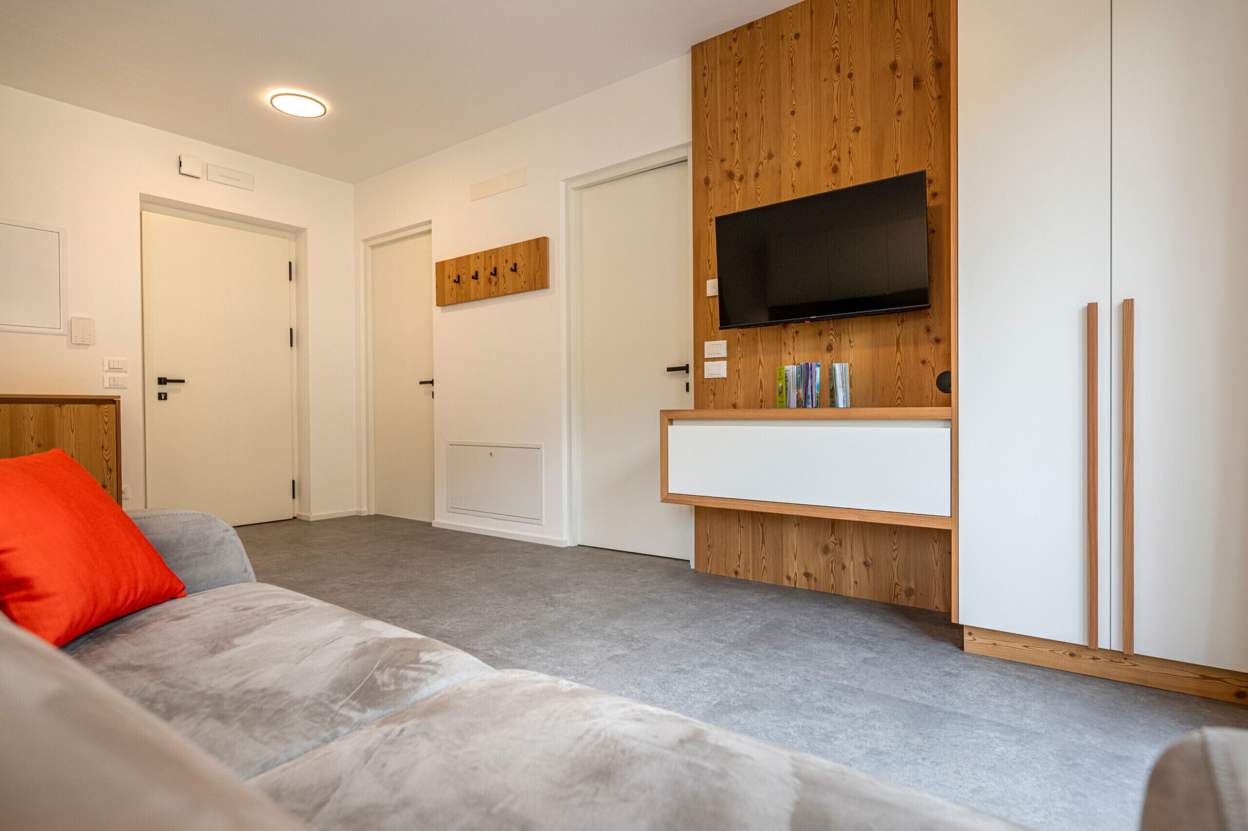 Un salon moderne avec un canapé gris, un coussin rouge, un téléviseur à écran plat fixé à un mur en bois et un meuble blanc placé en dessous.