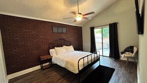 Un dormitorio con pared de ladrillo, una cama con ropa de cama blanca, un ventilador de techo, una mesita de noche y una silla cerca de un ventanal corredero.