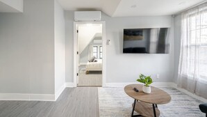 A modern living room with a flat-screen TV mounted on the wall, a round wooden coffee table with a plant, and a view into a bedroom through an open door. - 2 Bed Apt in BlackRock by beach (Bridgeport)