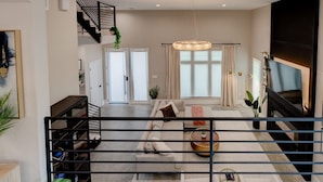 Una sala de estar moderna con un sofá, una mesa de centro y una televisión. Se observa una escalera con barandilla y una planta en la parte superior.