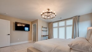 Un dormitorio con una cama grande, un televisor montado en la pared, una araña y una ventana con cortinas.