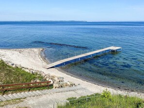 A coastal scene with a wooden pier extending into the water, a sandy beach, and a grassy area with a wooden fence.