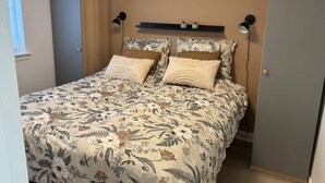 Ein Schlafzimmer mit einer blumigen Tagesdecke, zwei Nachttischlampen, einem Regal mit Dekoration und einer Tür links.