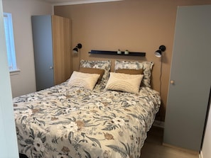Une chambre à coucher avec une couvre-lit à motifs floraux, deux lampes de chevet, une étagère avec des objets décoratifs et une porte sur le côté gauche.