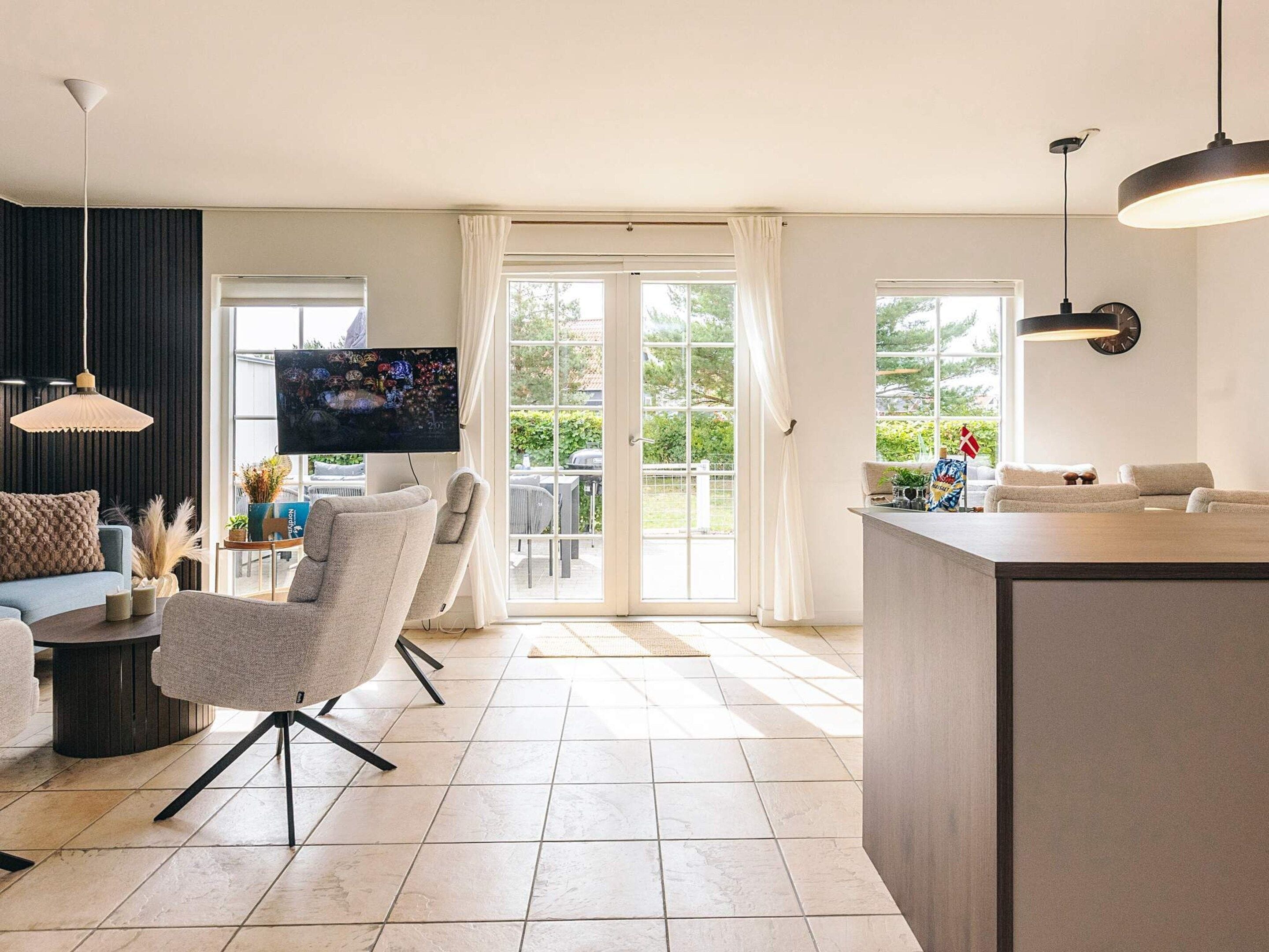Ein modernes Wohnzimmer mit Sofa, Sessel und Fernseher, ein Küchenbereich mit Bar sowie große Fenster mit Vorhängen.
