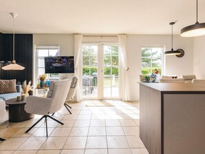 A modern living room with a sofa, armchair, and TV, a kitchen area with a bar, and large windows with curtains.
