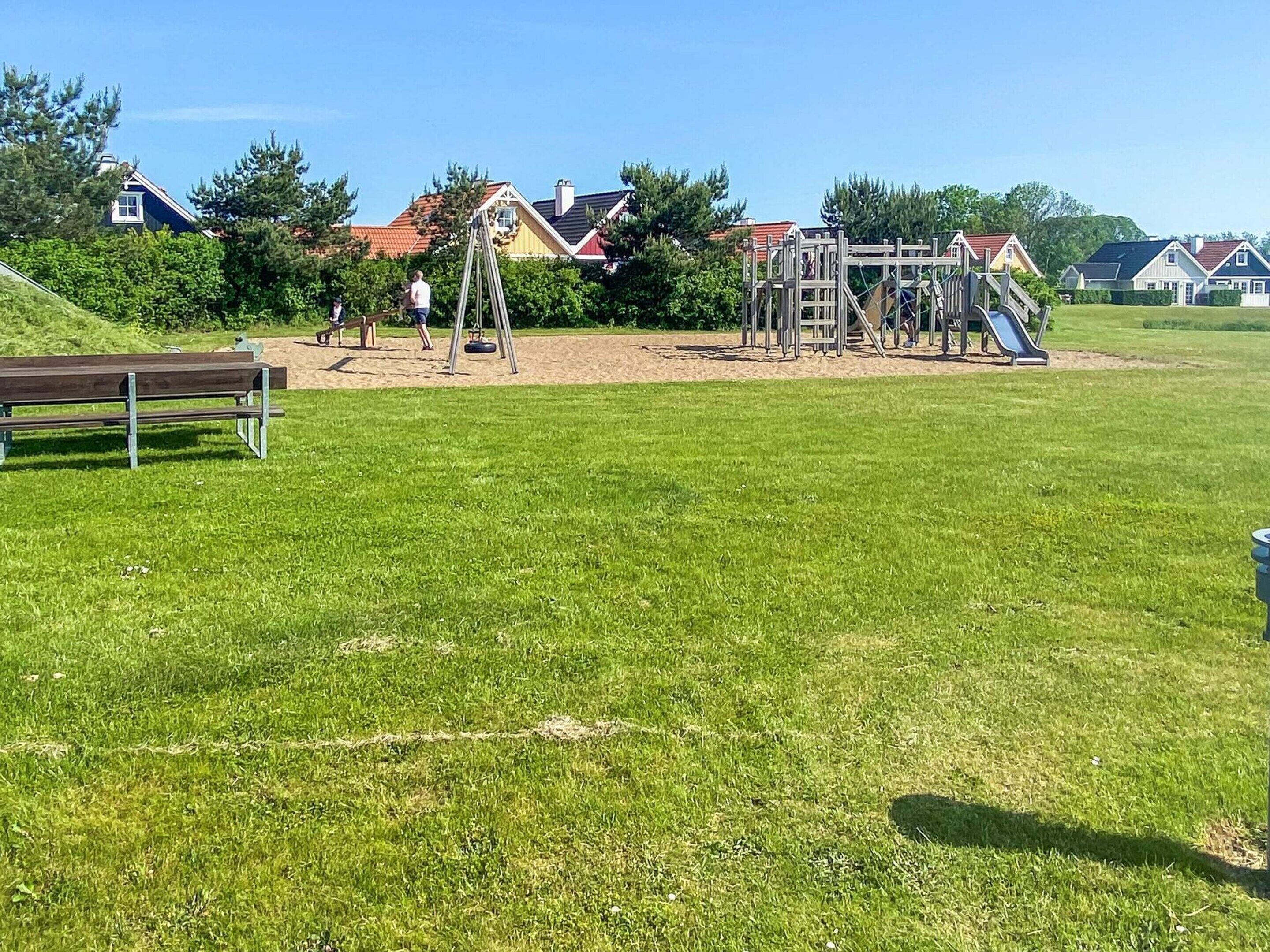 A playground with a swing set and climbing structures, surrounded by a grassy field and houses in the background.