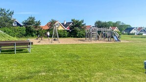 Ein Spielplatz mit Schaukeln und Klettergerüsten, umgeben von einer Wiese und Häusern im Hintergrund.