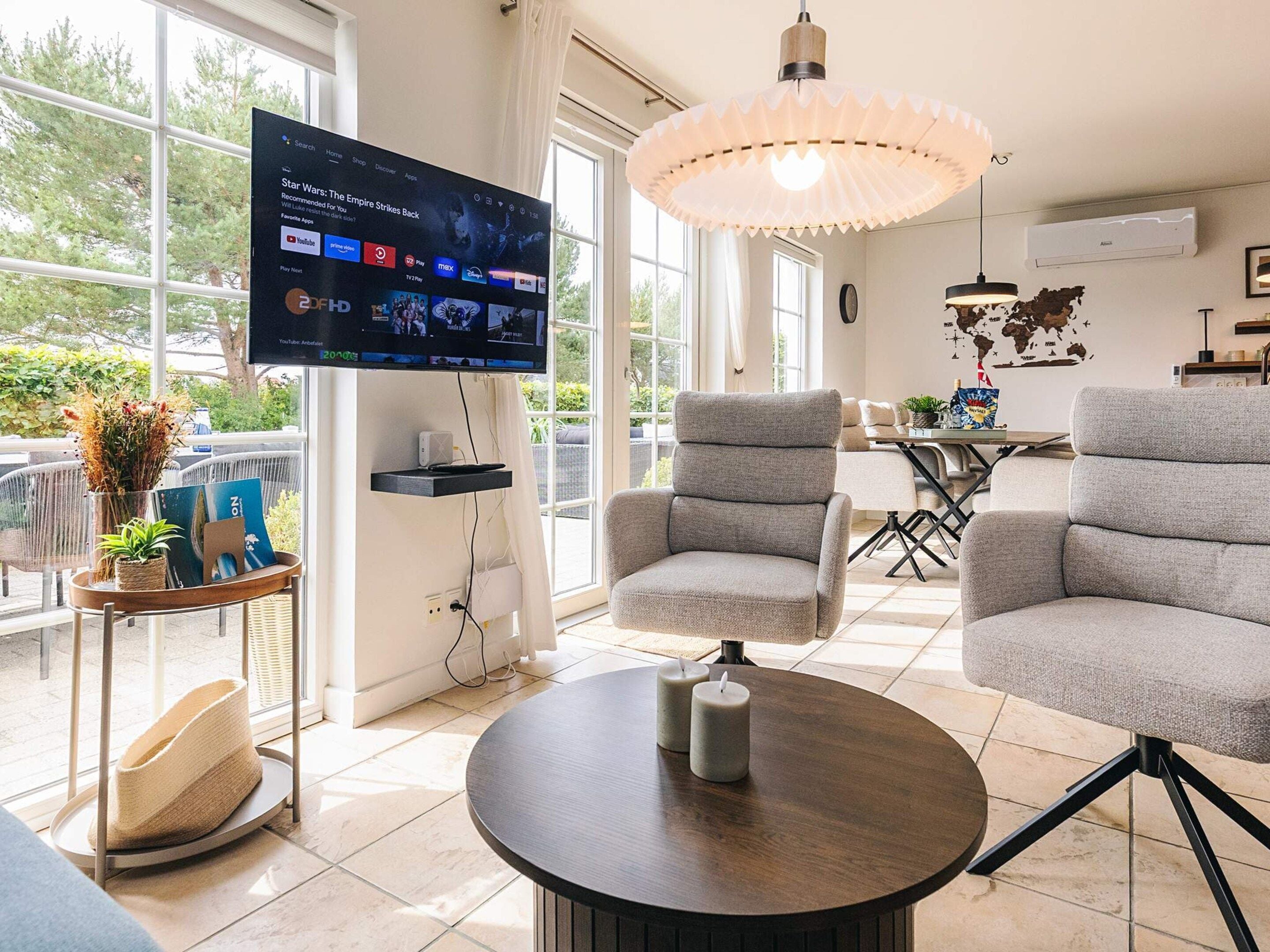 A modern living room with a flat-screen TV, grey armchairs, a round coffee table, and a decorative hanging light fixture.