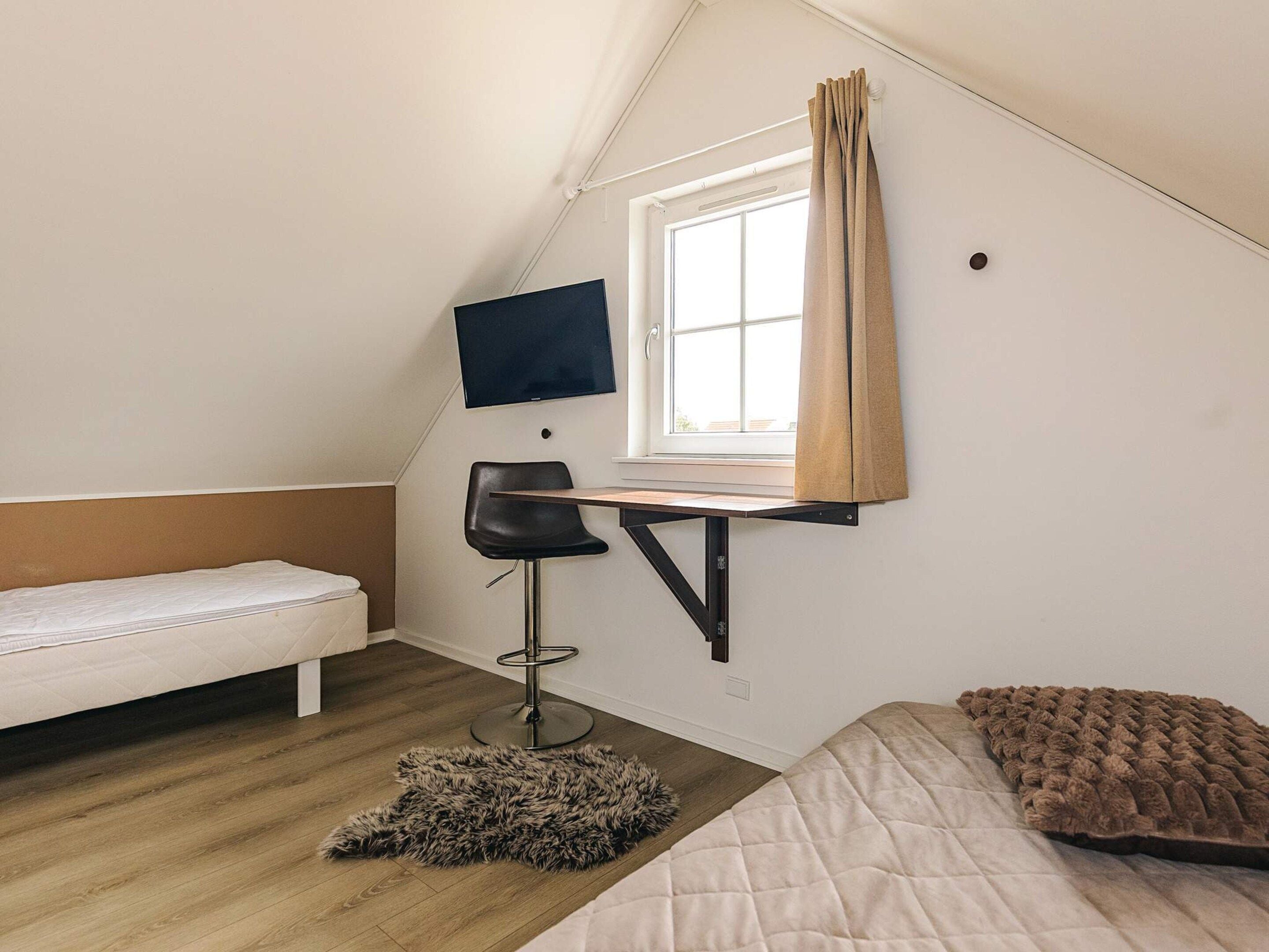 A small, triangular room with a bed, a desk with a chair, a television mounted on the wall, and a window with curtains.