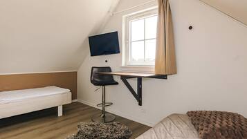 A small, triangular room with a bed, a desk with a chair, a television mounted on the wall, and a window with curtains.