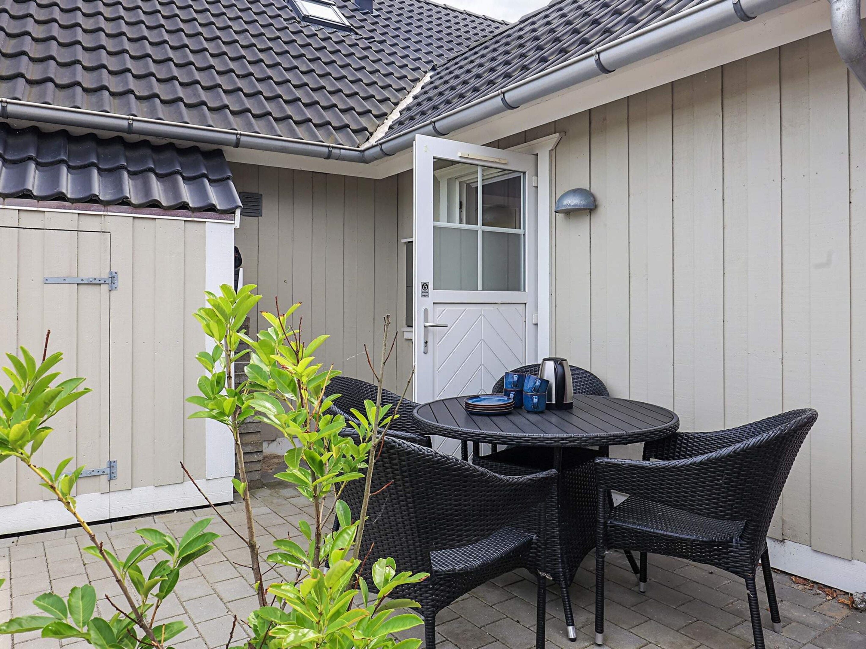 A patio area with a round table and four chairs, a door, and a small garden with green plants.