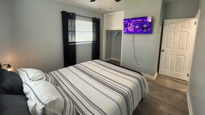 Un dormitorio con cama, ventilador de techo, ventana con persianas, televisor montado en la pared y una puerta.