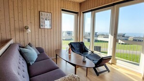 A modern living room with a wooden ceiling, a sofa, a chair, and large windows offering a view of the sea and buildings. - Well-maintained Holiday Home in Faborg (Faaborg)