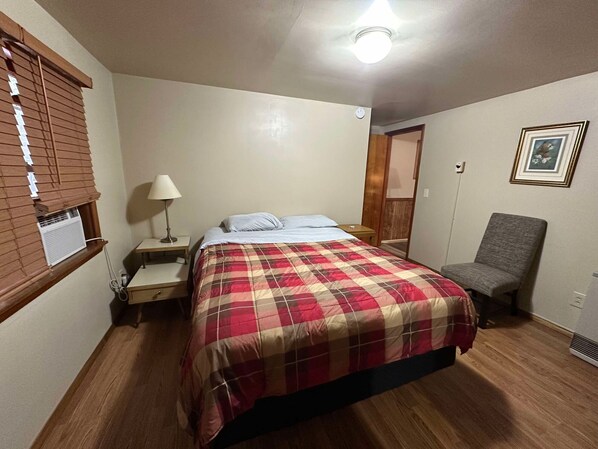 Un dormitorio con cama, una silla, una mesita de noche, una lámpara y una ventana con persianas.