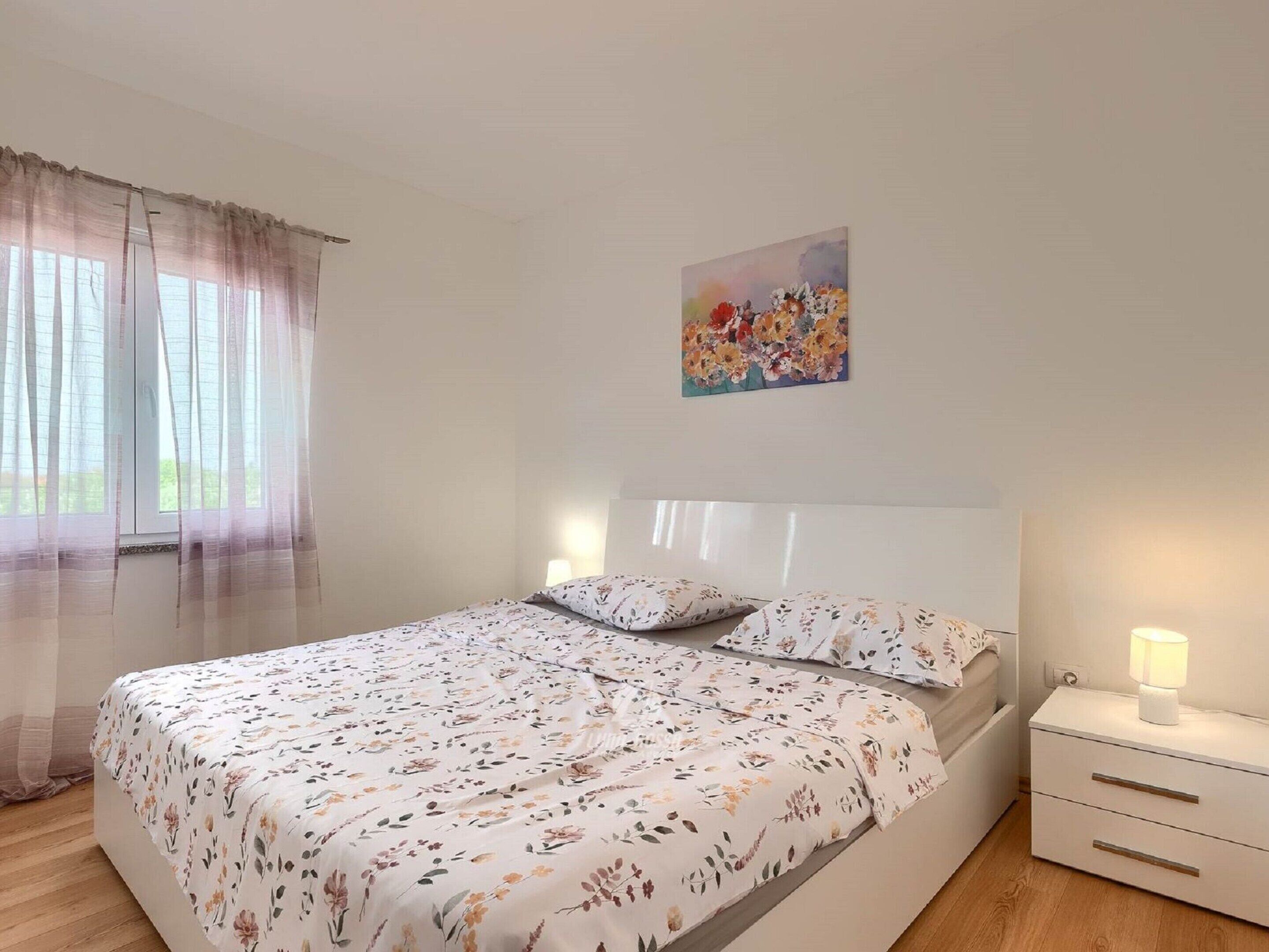 A bedroom with a bed, a nightstand, a lamp, and a window with sheer curtains.