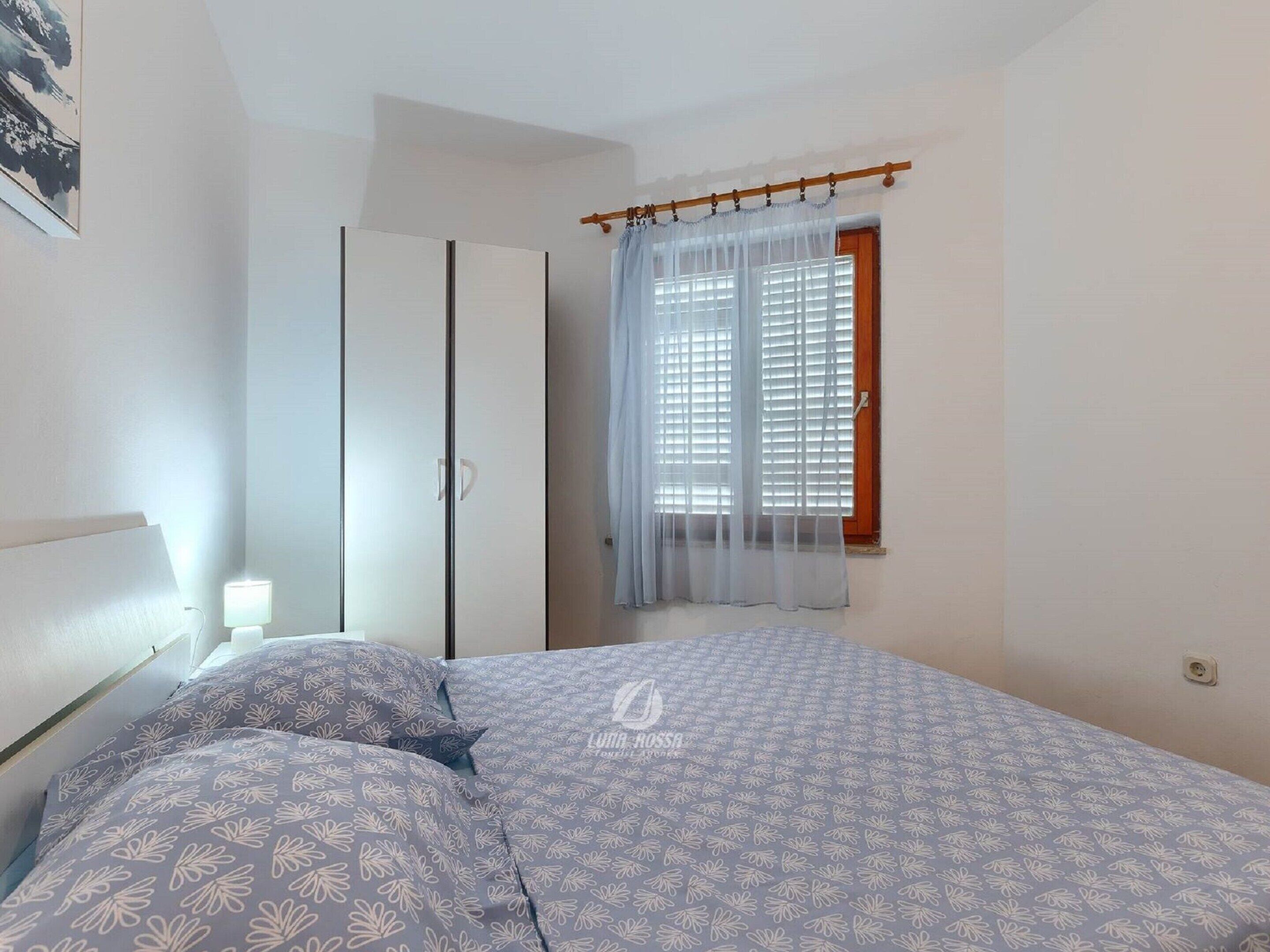 A bedroom with a bed, a wardrobe, a window with curtains, and a wall-mounted light.