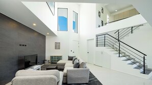 A modern living room with a sectional sofa, a flat-screen TV, and a fireplace. There is a staircase with a black railing leading to an upper level.
