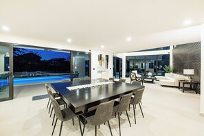 Een moderne eetruimte met een grote tafel en stoelen, uitzicht op een zwembad, en een gezellige zithoek met banken en een lamp.