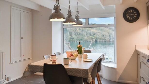 A dining area with a large table, chairs, hanging pendant lights, and a view of a lake through a window. - 3 bedroom accommodation in Fowey (Fowey)