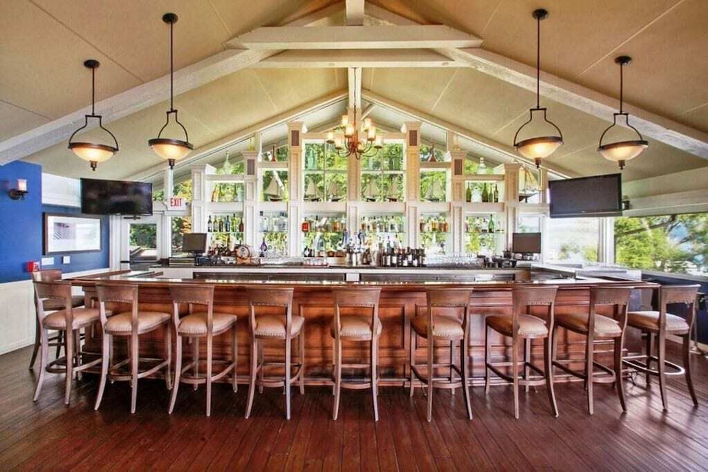 Un espace bar avec des tabourets de bar en bois, un plafond haut avec des poutres apparentes et une grande fenêtre offrant une vue sur la verdure environnante.