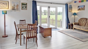 Ein Essbereich mit einem Holztisch und Stühlen, einem kleinen Beistelltisch und einem Sofa. Ein großes Fenster bietet Blick auf eine grüne Landschaft und einen weißen Zaun.