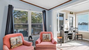 Una habitación con dos sillones rosados, una mesita, kitchenette y grandes ventanales con vistas al agua.
