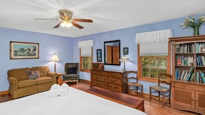 Un dormitorio con cama, cómoda de madera, estantería, ventilador de techo y ventana con vistas.