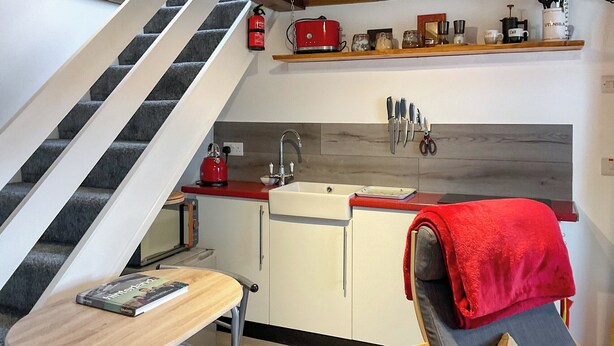 A compact kitchen with a red kettle, a sink, and a countertop. There is a chair with a red towel and a table with a book. A staircase is visible in the background. - 1 bedroom accommodation in Tregaron (Tregaron)
