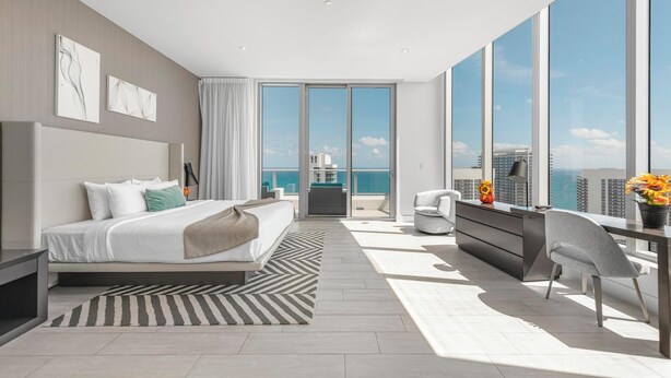 Une chambre moderne dotée d’un grand lit, d’un bureau et offrant une vue sur la ville grâce à de grandes fenêtres du sol au plafond.