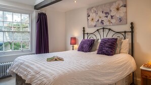 A bedroom with a large bed, a window with curtains, a radiator, and a floral painting on the wall.