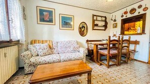 A cozy living room with a floral sofa, wooden dining table, and chairs, a patterned floor, and wall-mounted kitchen utensils. - Appartamento Maison Des Alpes - Affitti Brevi Italia (Sauze d´Oulx)