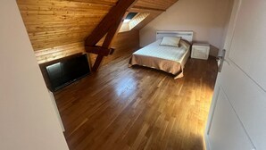 A bedroom with a sloped ceiling, wooden floor, a bed with a white headboard, a bedside table, and a wall-mounted TV. - Villa Le domaine des Rêves (Plateau d'Hauteville)