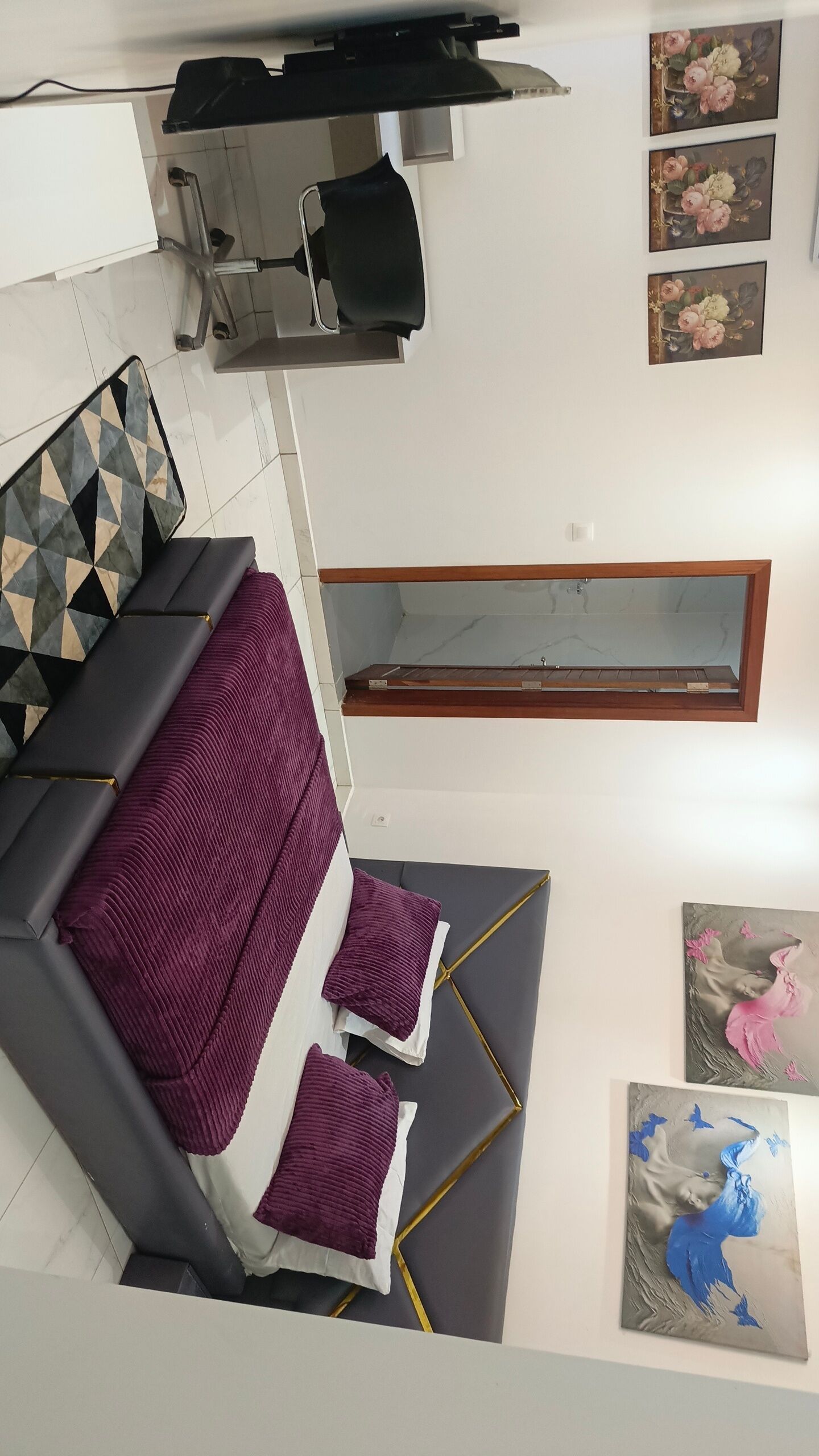 A modern living room with a sofa, a coffee table, and a TV. There are paintings on the wall and a window with a wooden frame.