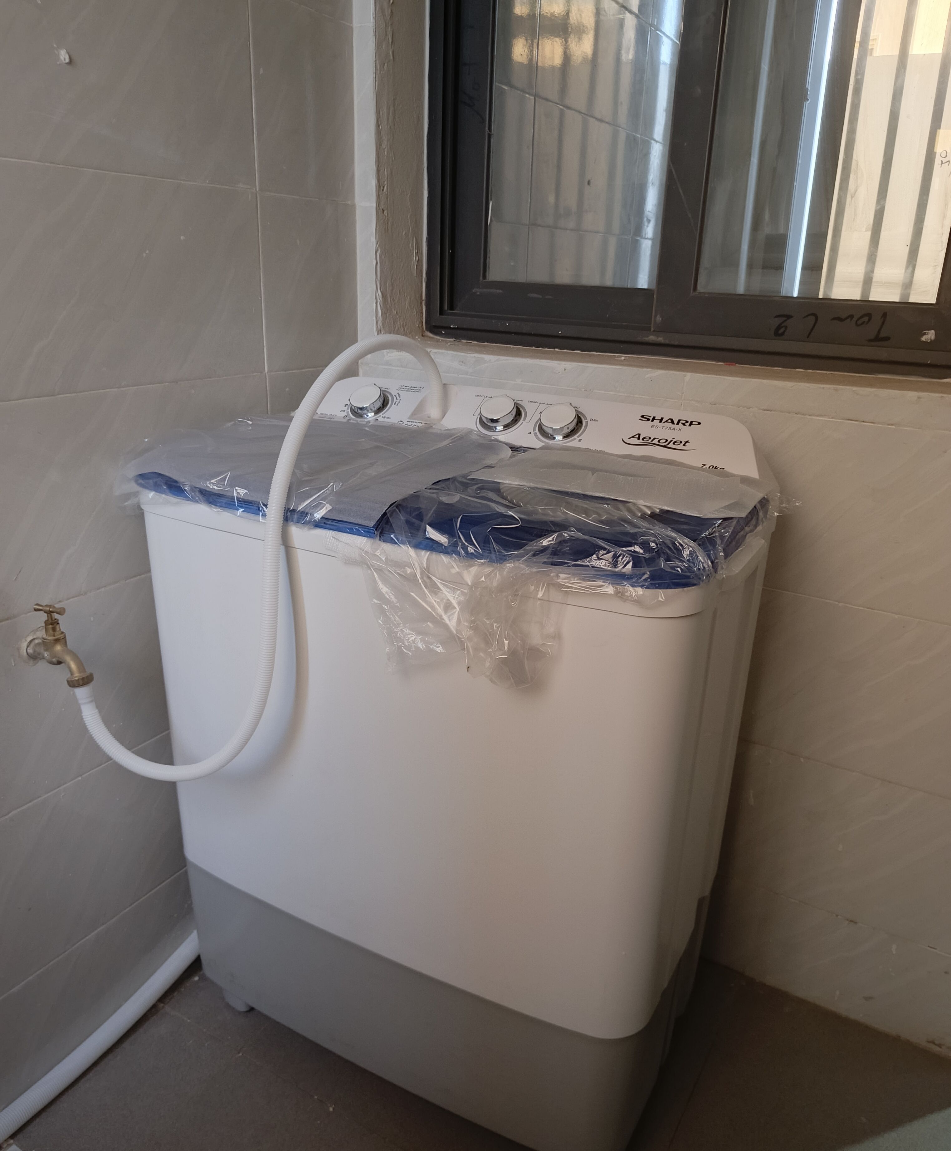 Un chauffe-eau compact, de couleur blanche, muni d’un tuyau, placé dans un coin avec des murs carrelés et une fenêtre au-dessus.