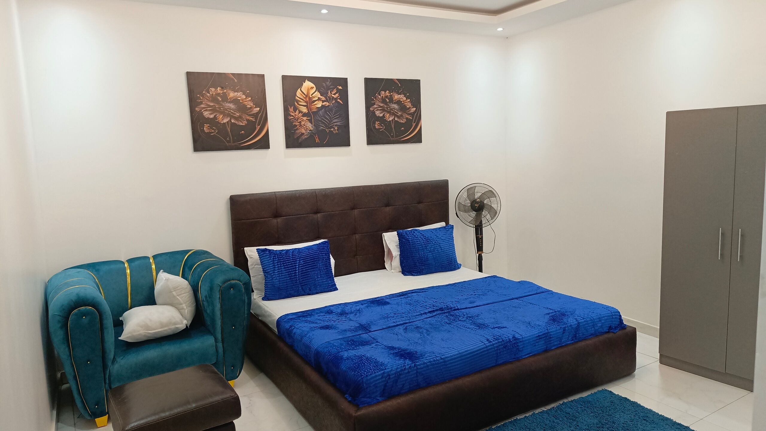A bedroom with a bed, a blue upholstered chair, a wardrobe, and three wall-mounted artworks.