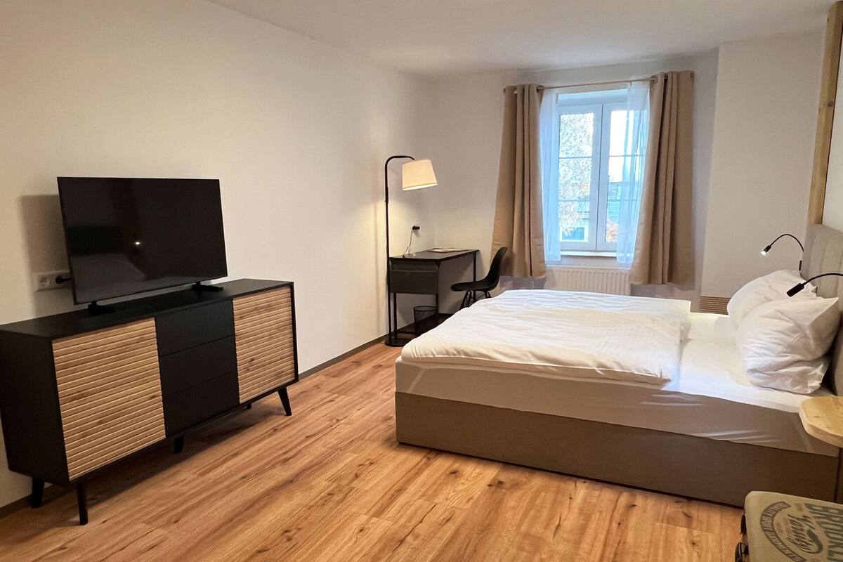 Quarto de hotel com cama, escrivaninha, cadeira, televisão e janela com cortinas.
