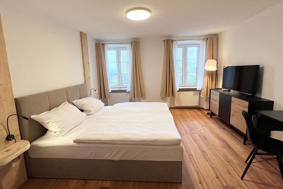 Quarto de hotel com uma cama grande, uma mesa com televisão e duas janelas com cortinas.