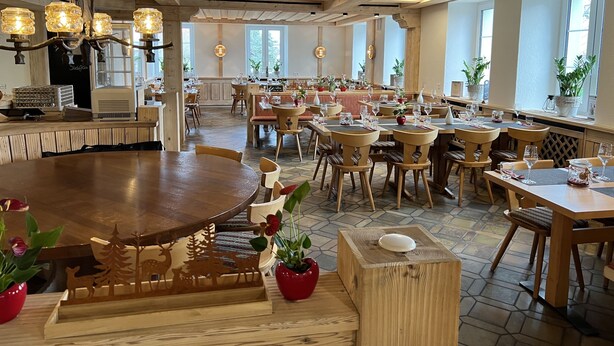 Een restaurant met houten tafels en stoelen, grote ramen en een hoge plafond met zichtbare balken.
